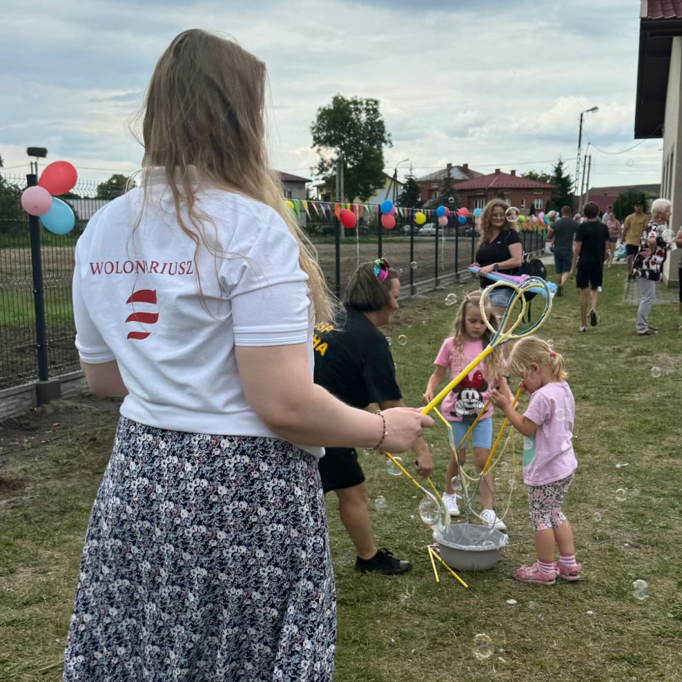 Młoda kobieta w koszulce z napisem Wolontariusz z logotypem PFR puszcza bańki wraz z dziećmi